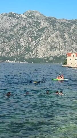 A vertical outdoor shot captures a scenic view of a bay with people swimming and kayaking. In the foreground, approximately eight people are seen swimming in the clear blue water. Further back, two individuals are in a green kayak, with one person paddling. The water has a slight ripple and appears transparent near the shore, revealing the seabed. On the right side of the frame, a traditional-style building with a red tile roof and light-colored walls stands by the water's edge. In the background, a majestic, rugged mountain range dominates the landscape under a clear blue sky. A few distant boats or buoys are visible on the water's surface towards the left. The overall scene suggests a pleasant day at a coastal location.