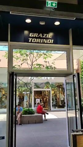 The video shows an outdoor shopping area. In the foreground, there are shelves displaying children's shoes, specifically sandals and sneakers. The shoes are colorful, with pink, purple, and teal as dominant colors. There are also shoe boxes stacked beneath the shelves. Through an open doorway, the scene continues outdoors. Above the doorway, a sign reads "GRAZIE TORINO!" in large yellow letters on a dark blue background. A tree with green leaves is visible through a large window above the doorway. Beyond the doorway, in the outdoor area, two people are sitting on a bench. One person, wearing a red shirt, is facing away from the camera. Another person, wearing a blue shirt, is also sitting on the bench, facing left and looking down. Further in the background, several people are walking. One person is carrying a large blue bag. Stores are visible on either side of the walkway, with signs that are partially obscured. A tall green plant is positioned to the right of the doorway. The overall environment appears to be a modern outdoor mall or outlet shopping center on a sunny day.