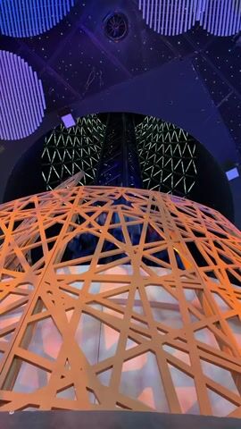 This media file captures a low-angle shot from within an architectural structure, looking upwards. The foreground is dominated by a large, intricate, geometric dome made of light-colored wooden slats, creating a lattice-like pattern. Behind this dome, a tall, dark, slender tower rises towards the ceiling. The tower appears to be made of metal beams and is illuminated with blue light. The background features a large, dark, vaulted ceiling adorned with a complex geometric pattern of illuminated lines, creating a starry or crystalline effect. Circular, striped light fixtures in shades of blue and purple are also visible on the ceiling. The overall lighting suggests an indoor, possibly nighttime or dimly lit environment. The scene appears to be part of a modern exhibition space or architectural installation, likely in a public area. There is no discernible activity or people visible, focusing solely on the architectural elements. The context provided places this scene in Doha, Qatar, suggesting it might be part of a pavilion or building at an expo or cultural landmark. The ambiance conveyed is one of awe and wonder, emphasizing the scale and design of the structure.