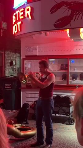 A young man with brown hair wearing a dark red tank top and blue jeans plays a brass instrument resembling a French horn. He is standing on a cobblestone surface outside a building with a neon lobster sign that reads "LOBSTER HOT." A black dog sits to his left. In front of him is a large brass instrument, possibly a tuba or sousaphone, coiled on the ground. The building has white siding and red trim. A trash can is visible on the left. The scene is illuminated by red neon lights and the ambient light of the evening. There are several people in the foreground, partially visible, suggesting an audience. The overall impression is of a street performance.