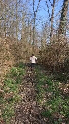 A young person walks down a muddy path in a forest. The person is wearing a light-colored jacket, dark pants, and has dark hair in a ponytail. The trees surrounding the path are mostly bare, with some brush and green grass visible. The sky is blue and clear.