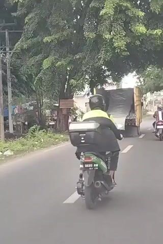 A video shows a person riding a green motorcycle on a road. The rider is wearing a yellow and green jacket and a black helmet. The motorcycle has a black storage box on the back. There is a large truck in front of the motorcycle, also traveling on the road. The truck is covered with a tarp. Other motorcycles are visible on the road as well, further ahead. The road is paved and lined with trees. The scene appears to be in a semi-urban or suburban area.