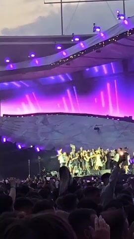 A crowd of people are gathered at an outdoor concert or event. A large stage with purple and pink lights is visible in the background, with a group of people standing on it. The audience is densely packed, with many individuals holding up their phones to record the event. The overall atmosphere appears to be energetic and celebratory.
