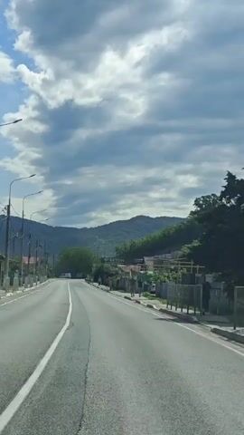 The video depicts a straight, paved road with a solid white line marking the shoulder. Street lights line the left side of the road, supported by poles. On the right, there are houses behind a fence, with a sign that reads "БЕТОН". In the distance, a mountain range covered in green trees stretches across the horizon. The sky is blue with scattered white and gray clouds, some of which are obscuring the view of the mountains. A few people can be seen walking on the sidewalk near the houses, and a few cars are visible in the distance. The camera appears to be mounted inside a moving vehicle.