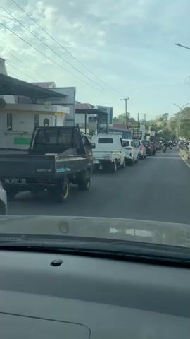 The video shows a line of vehicles, mostly cars and trucks, stopped on a road. The camera is inside a vehicle, looking forward. The sky is cloudy, and there are buildings and trees visible on the side of the road. Some people are standing near the vehicles.