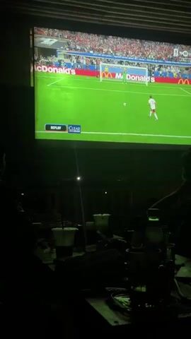 The video shows a large projector screen displaying a soccer game. The game appears to be a penalty kick, with a player in a white and red uniform lined up to take the shot. The goal is visible, with a goalkeeper in yellow. There are advertisements for McDonald's around the field. In the foreground, there are several people sitting at a table, watching the game. Drinks and other items are on the table. The room is dimly lit, with the light from the projector illuminating the screen. The overall atmosphere appears to be relaxed and casual, likely a bar or lounge setting.