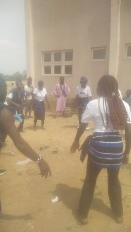 A group of people are dancing outside a beige building. They are dressed in coordinated outfits, some with blue and white striped fabric. One person has long brown braids. The ground is covered in tan dirt and some scattered debris. The video is shot from a low angle with the camera turned on its side.