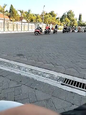 A group of motorcycles are moving in a procession on a paved road. The motorcycles are moving from left to right in the frame. The road is made of dark gray pavers. There is a curb and a drainage grate on the right side of the road in the foreground. In the background, there are trees, buildings, and a fence. The sky is blue and there is bright sunlight. The motorcycles appear to be in motion, and the people on them are wearing helmets.
