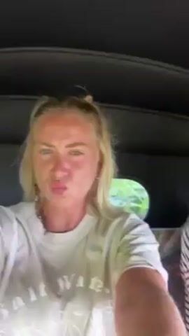 Two women are inside a vehicle, likely a tuk-tuk or similar open-air taxi. The woman on the left has blonde hair, a white t-shirt, and is looking at the camera. The woman on the right has her blonde hair in a ponytail, and is wearing a striped shirt. The vehicle's interior is visible, and the background shows glimpses of trees and vegetation. The video appears to be taken from the perspective of the woman on the left.