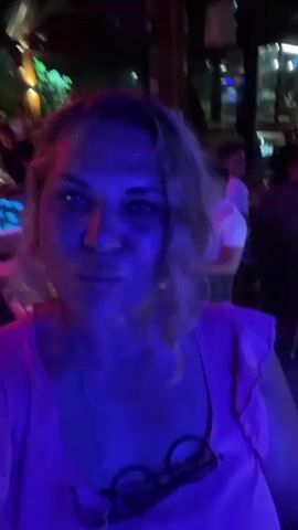 A woman with blonde, wavy hair is filming a selfie video in a dimly lit bar or restaurant. She is wearing a pink top and has a pair of glasses tucked into the neckline. The background is blurred but shows people sitting at tables and a band performing on a stage with blue and red lights. The woman looks directly at the camera with a playful expression.