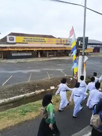 The video shows a group of young people in white uniforms marching in formation on a paved area. They are wearing black hats, and some have white gloves. The marching group is accompanied by two people in darker clothing, one of whom appears to be holding a bag. In the background, there is a yellow building with signage that includes the words "PAVILION OLEH-OLEH & JAJANAN PARAHYANGAN". There are also several flags visible, including an Indonesian flag. The ground is wet, suggesting it may have recently rained. The overall scene appears to be an outdoor event or parade.