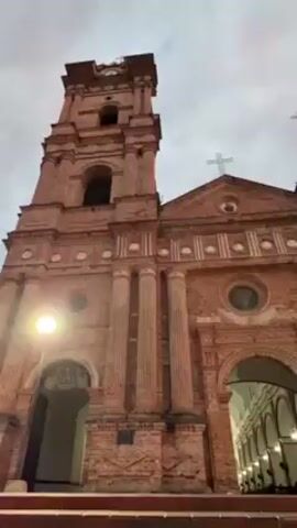 The video showcases a low-angle view of a large, brick church with a prominent bell tower, set against a dramatic sky. The church features classical architectural elements such as arches, columns, and a central dome with a cross on top. The brickwork shows signs of age and weathering. The sky is a mix of gray and pink hues, suggesting either sunrise or sunset, with distinct cloud formations. There is a glimpse of streetlights and distant hills in the background. No people are visible in the frame. The camera smoothly pans upwards, focusing on the sky and the church’s facade.