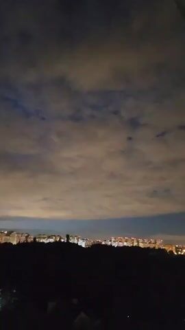 The video shows a nighttime cityscape viewed from an elevated position. In the foreground, silhouetted trees and foliage create a dark, shadowy border. Beyond this, a strip of buildings with illuminated windows stretches across the midground, indicating an urban area. Above the city, the sky is filled with dense, textured clouds, illuminated from below by the city lights, giving them an eerie, yellowish glow. Patches of darker blue sky are visible through gaps in the clouds. The overall impression is of a tranquil, albeit slightly dramatic, night scene. There are no people or specific actions depicted.