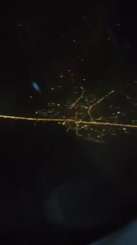The video shows a nighttime aerial view of city lights and roadways. The camera pans and zooms, focusing on the illuminated grid of streets and buildings. In some segments, the lights appear as blurred streaks, suggesting movement or a long exposure. There are also instances of what appear to be reflections or artifacts on the camera lens.