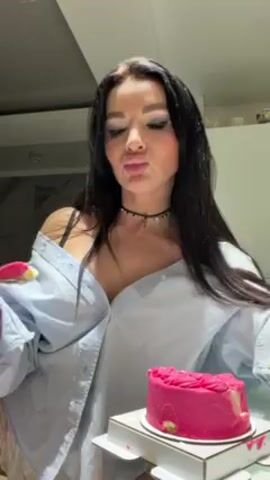 A woman is eating a small pink cake, possibly in a kitchen or dining area. She is wearing a light blue button-up shirt that is unbuttoned, showing some cleavage. She has long, dark hair, and is wearing a choker. She has gotten some of the cake on her nose. The cake sits in a cardboard box on a table.