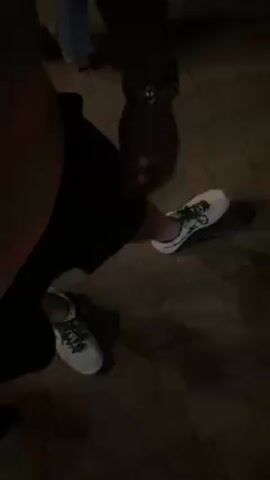 Here is a concise description of the video:

The video shows a person holding an iPhone displaying the time as 22:45, indicating 10:45 PM. The phone's wallpaper is colorful and patterned. The person is wearing a gold watch. On the ground nearby are two white sneakers. The location appears to be outdoors, possibly on a paved area. There are other people visible in the background.