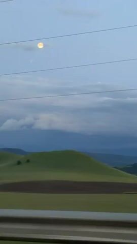 The video shows a landscape scene with rolling green hills under a cloudy sky. Power lines are visible in the foreground, stretching across the frame. The moon is visible in the upper portion of the sky. The camera appears to be in motion, possibly from a vehicle, as the landscape shifts from left to right. The lighting suggests it is either early morning or late evening. There are scattered trees on the hills and patches of brown earth, indicating cultivated fields. No people or animals are visible in the video.