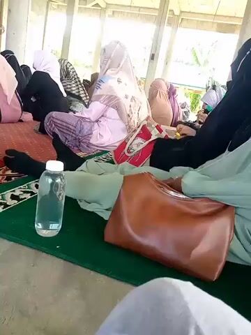 A group of women are seated on prayer mats in an outdoor covered area. They are all wearing hijabs and modest clothing. One woman in the foreground is wearing a light green hijab and holding a brown purse. To her left is a clear plastic bottle filled with water. Behind her, another woman is holding a cardboard box with the words "KOTAK INFAQ" written on it in large black letters. Several other women are visible in the background, also seated on prayer mats. The environment appears to be a communal space, possibly a mosque or a prayer hall, with an open-air design and visible support beams. The lighting suggests it is daytime.