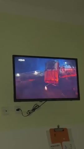 A flat-screen television mounted on a pale green wall displays a scene from what appears to be a movie or television show. The screen shows a nighttime cityscape with a bridge illuminated by streetlights and glowing with fire and explosions. A large vehicle, possibly a truck, is visible on the right side of the screen, its rear end illuminated by the intense orange glow of the fires. The logo "nova" is visible in the top left corner of the screen. Below the TV, a white electrical outlet is mounted on the wall. A black power cord runs from the TV to the outlet. A clipboard with papers is hanging on the wall below the television. The overall lighting in the room is dim, suggesting it is nighttime or the lights are turned off.