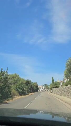 A POV shot from a vehicle moving down a paved road. The road curves gently to the right, with trees lining the left side of the road and a stone wall on the right. In the distance, several white buildings with domed roofs are visible on a hill. The sky is a clear blue with a few wispy white clouds.