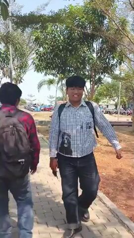 Two young men are walking down a paved path outdoors. The first man, facing the camera, is wearing a checkered shirt and jeans. The second man is walking away from the camera, wearing a black jacket and backpack. In the background, there are trees, a building with windows, and a blue structure with barrels. The scene appears to be a campus or park.