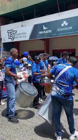 The video shows a group of male drummers performing outdoors. The drummers are wearing blue shirts and jeans. They are playing a variety of drums, including snare drums and bass drums. A crowd of people, including children in school uniforms, are watching them. There is a building behind the drummers with signs that read "FERRETERIA RIO TUY, C.A.", "PRO-PLASTIC", "ECO-COLOR", and "COLOR FRUT". The scene appears to be part of a parade or celebration.