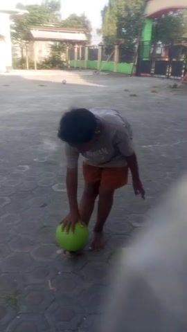 A young boy is playing with a bright green ball in an outdoor paved area. He is barefoot and wearing a grey t-shirt and orange shorts. The boy is shown kicking the ball into the air and then bending down to retrieve it. In the background, there are buildings, trees, and a fence. The lighting suggests it is daytime.
