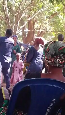 The video shows a group of people dancing outside under a tree. The view is from the back of a person sitting in a blue plastic chair. There are at least five other people visible, most of them women wearing colorful headscarves. A man in a dark blue shirt is dancing with his arms raised. A young girl in a pink dress stands nearby. The environment seems to be a yard with trees providing shade. The overall atmosphere appears lively and celebratory.