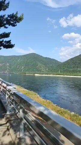 This video captures a scenic view of a wide, flowing river with lush, tree-covered mountains in the background. The sky is bright blue with scattered white clouds, suggesting a pleasant, possibly sunny day. The riverbanks are lined with grass and rocks, and a white railing runs along the bottom of the frame, indicating a viewpoint from a bridge, walkway, or roadside.

In the middle ground, a large hydroelectric power station complex is visible on the far bank, nestled against the mountainside. The imposing structure of the dam and associated buildings suggests industrial activity within this natural landscape. The water is dark and appears to be in motion, with subtle ripples and reflections.

The overall impression is one of natural beauty juxtaposed with human engineering. The time of day appears to be daytime, likely late morning or afternoon, given the brightness and the quality of light. There is no visible text within the video. The location context of Cheremushki, Russia, is consistent with the presence of a large river and mountainous terrain, and is known for the Sayano-Shushenskaya Dam, which the visible structure strongly resembles. The scene evokes a sense of vastness and the power of both nature and technology.