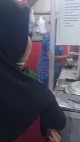 Here is a concise description of the video:

The video shows the side view of a woman wearing a black hijab standing in front of a food stall. Another woman, wearing a blue floral dress, is behind the food stall, preparing food. A sign listing menu items is visible on the side of the stall. The area around the stall appears to be a simple, possibly informal, food preparation and service space.
