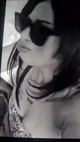 A low-angle, black-and-white video shows a woman with dark hair wearing sunglasses and a tube top. She is facing forward and slightly to the left. A man in a suit is visible in the background. The video appears to be taken from inside a car.