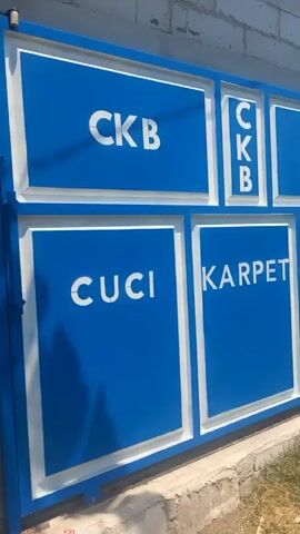 The video shows a blue metal gate with white lettering. The gate is divided into several rectangular panels. The top row of panels has the letters "CKB" in each. The bottom row of panels has the words "CUCI", "KARPET", and "BANGIL" from left to right. A white brick wall is visible behind the gate, and some green foliage is growing along the bottom of the gate. The lighting suggests it is daytime.