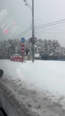 The video depicts a snowy scene where a person is climbing a ladder attached to a utility pole. The person is wearing dark clothing and a backpack, and they are ascending the ladder in a snowy environment. The utility pole is positioned next to a road sign with a blue and white arrow, and another red and white chevron sign is nearby. In the background, there are snow-covered trees and a sign with the word "MOCKBA" visible. The ground is entirely covered in snow, and the weather appears to be overcast. The video is taken from inside a vehicle, looking out onto the scene.