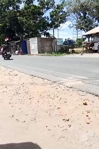 A motorcycle carrying a person is traveling on a paved road. The motorcycle is seen from the side. The person on the motorcycle appears to be a driver. In the background, there are several small structures, trees, and power lines. The area appears to be rural or semi-urban with a dirt shoulder on the side of the road. The sun is shining.