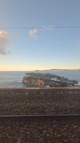 The camera captures a side view from a moving train, revealing train tracks and a stone wall running parallel to the sea. Beyond the wall, the calm, blue sea extends to the horizon. A small, rocky, tree-covered island is situated near the shoreline, and a distant landmass is visible on the horizon. The sky is a clear blue with scattered white clouds, and the sun appears to be setting. The scene is peaceful and serene.