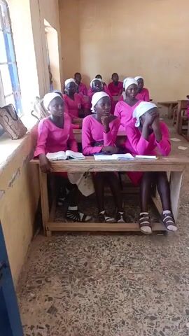 The video shows students in a classroom. The students are young women and girls, all wearing pink dresses and white headscarves. They are seated at wooden desks, some with open books or papers in front of them. Some are resting their heads on their hands, looking at the camera. The room is brightly lit, and the walls are plain. There is a window on the left side of the frame. The floor is covered with speckled tile. The scene is still and focused on the students.