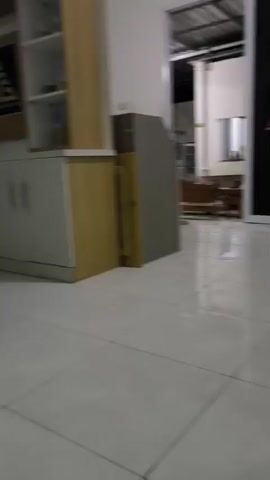 The video shows a blurred interior scene. In the foreground, a white tiled floor is visible, with faint shadows cast by unseen objects. To the left, a white wall creates a corner. Further back, a large piece of furniture with a light wood and white finish is against the wall. It appears to be a cabinet with drawers or doors, and possibly shelves above. There is a dark framed picture or panel above the lower cabinets. To the right of this furniture, a white door with a dark glass panel is visible. Beyond that, another similar white door with a dark glass panel is slightly further away. An illuminated green sign with white text is mounted above the second door. A white pillar is also present in the right side of the frame, partially obscuring the view of the wall and furniture to its right. The overall impression is of a hallway or a room with minimal furniture and a clean, modern aesthetic.