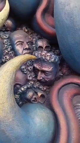 The video is a close-up, slow pan across a relief sculpture. The sculpture depicts multiple human faces and numerous small skulls embedded in a dark, textured background. The faces appear to be male and are rendered with varying degrees of detail, some with distinct features and others more abstract. The skulls are smaller and more uniform in appearance. The sculpture is accented with curved, organic shapes in shades of blue, pink, and gold, which frame and overlap the faces and skulls. The lighting casts shadows that emphasize the depth and texture of the relief.