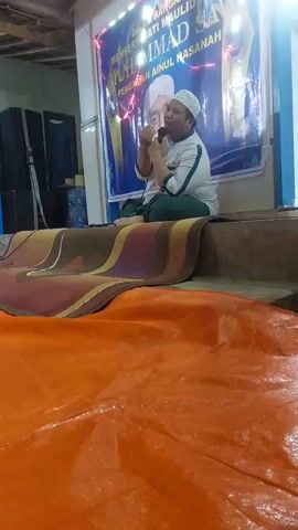 The video shows a man speaking into a microphone while seated on a stage. He is wearing a white cap, a green and white striped shirt, and green pants. Behind him is a banner with text that appears to be in Indonesian. The stage is covered with rugs, and in the foreground, there is a large orange tarp. To the left of the stage, there are two black speakers. The lighting suggests an indoor setting, possibly a community hall or a religious gathering. The man is gesturing with his hands as he speaks.