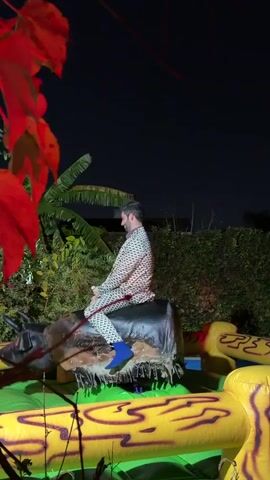 At night, a man rides a mechanical bull surrounded by a yellow and green inflatable structure. He is wearing a white and black patterned suit, a dark red shirt, and blue socks. He faces away from the camera and then turns to look and smile. The bull is placed in an outdoor area, with vegetation visible around it. The lighting changes from blue to red to green at different points during the video.