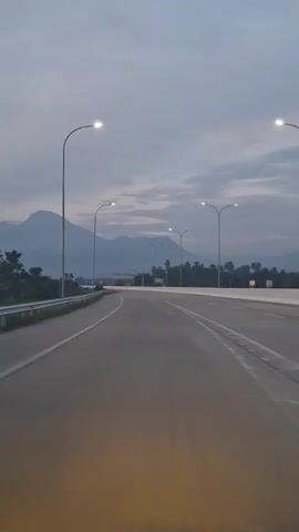The video shows a view from a vehicle driving on an empty highway. The sky is cloudy and appears to be either dusk or dawn. Tall, thin streetlights line the sides of the road, and their lights are on. In the distance, mountains are visible. The road has multiple lanes and is in good condition. The vehicle appears to be moving at a steady pace, and there are no other cars or obstacles in sight. The overall scene is calm and serene.