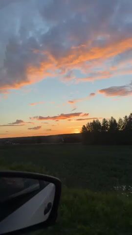 A car's side mirror is visible in the foreground of the video, reflecting a sunset. The sky is filled with dramatic clouds painted in shades of orange, pink, and blue. The sun is setting on the horizon, casting a warm glow over a landscape of green fields and trees. The video appears to be shot from inside a moving vehicle, as evidenced by the slight blur and the perspective of the side mirror.