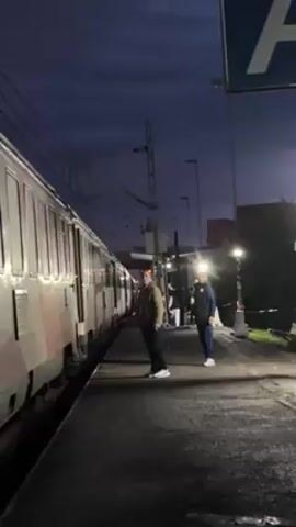 The video shows a train station platform at night. Two men are standing near a train, which is stopped alongside the platform. The man in the foreground is wearing a light-colored jacket, dark pants, and white shoes. The other man is wearing a dark tracksuit with white shoes. A sign with the letter "A" is visible in the upper right corner of the frame. Several light posts illuminate the platform. Overhead electrical wires are visible above the train. The sky is dark, suggesting it is either early morning or late evening.