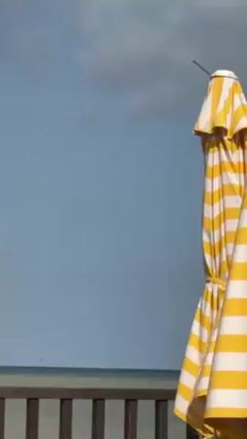 The video shows a beach scene. In the foreground, a brown wooden railing partially obscures the view of the water. To the right of the frame, a closed yellow and white striped beach umbrella stands tall. The sky above is blue with a few scattered clouds. The ocean is visible in the distance.