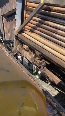 The video shows a machine processing long, cylindrical pieces of wood. The machine is yellow and has a system of chains and gears to move the wood pieces. The wood pieces are stacked horizontally in a metal frame attached to the machine. Water is being sprayed onto the wood as it moves through the machine. The machine is located outside near a wooden building. The video is shot from a high angle, looking down at the machine.