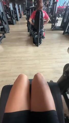 The video begins with a close-up shot of a person's legs on a gym machine, specifically a leg curl machine. The camera then pans up to show the person's torso and shoulders, and a water bottle is attached to the side of the machine. The camera then tilts up to reveal more of the gym environment, showing rows of exercise equipment and other people working out. In the background, a woman in a red shirt is using a similar leg machine. The video then returns to the initial shot of the person's legs on the leg curl machine, with the padded bar resting on their lower calves. The camera then focuses on the legs again as the machine appears to be in motion, though no actual movement is visible in the still frames. The scene is set in a well-lit, modern gym with wooden flooring and various weight machines.