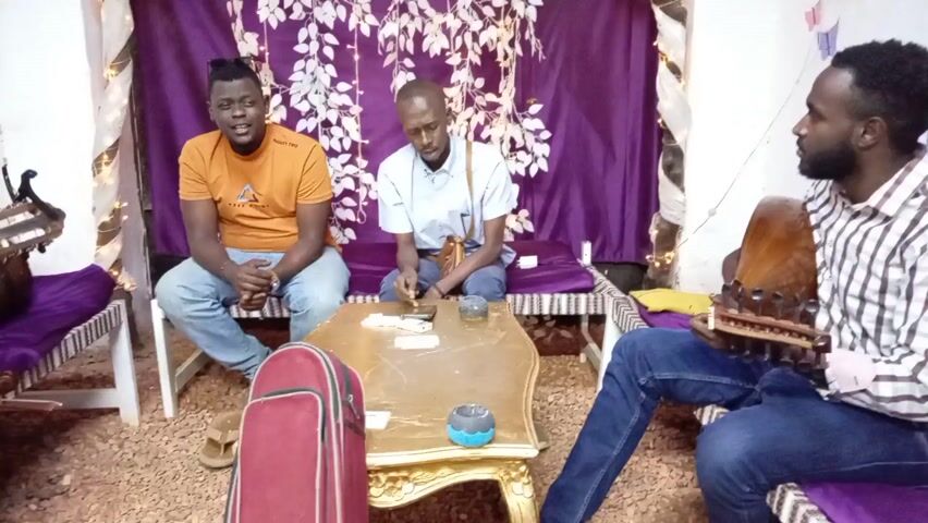 Three men are sitting in a decorated room and playing musical instruments. The man on the right is playing a stringed instrument that is shaped like a lute. The man in the middle is seated next to a small table with objects on it. The man on the left is playing a guitar. The room is decorated with a purple curtain, white floral decorations, and string lights. There is a red guitar case at the foot of the table.