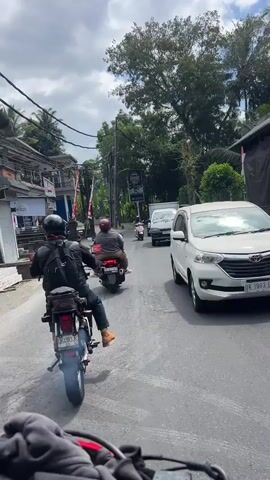 The video shows a street scene from the perspective of someone riding a motorcycle. In the foreground, a black motorcycle with a rider wearing a backpack and helmet is visible. The road is asphalt and appears to be cracked in places. To the left, there are buildings and storefronts. Further down the road, there are other motorcycles and vehicles, including a white van and a white car. Trees and foliage are abundant on both sides of the street, and the sky is cloudy. The overall impression is a busy street in a tropical or subtropical environment.