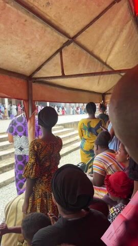 From under a covered structure, a scene unfolds showing a reenactment of the crucifixion of Jesus Christ. A person portraying Jesus is shown being crucified on a wooden cross. Other people are participating in the scene, and spectators are watching the performance. The location appears to be an outdoor setting, possibly a church or religious ground.