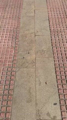 The video shows a paved area with a central pathway made of light-colored stone. On either side of this pathway are sections of red brick paving with a grid pattern. As the camera moves forward, the pathway leads to a circular garden bed with grass and various shrubs and trees. In the center of the garden is a bronze statue of a man holding a tool or object over his shoulder. Behind the garden, there are buildings, trees, and parked vehicles. The lighting suggests it is daytime.